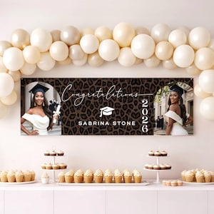May include: A graduation banner with a leopard print background and the words "Congratulations Sabrina Stone 2026". The banner features two photos of a graduate in a cap and gown, with a balloon arch above and cupcakes below.