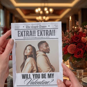 May include: A newspaper with the headline "EXTRA!! EXTRA!!" and the text "WILL YOU BE MY Valentine?" is held in front of a floral arrangement. The newspaper features a photo of a couple. The scene is set on a table with candles.