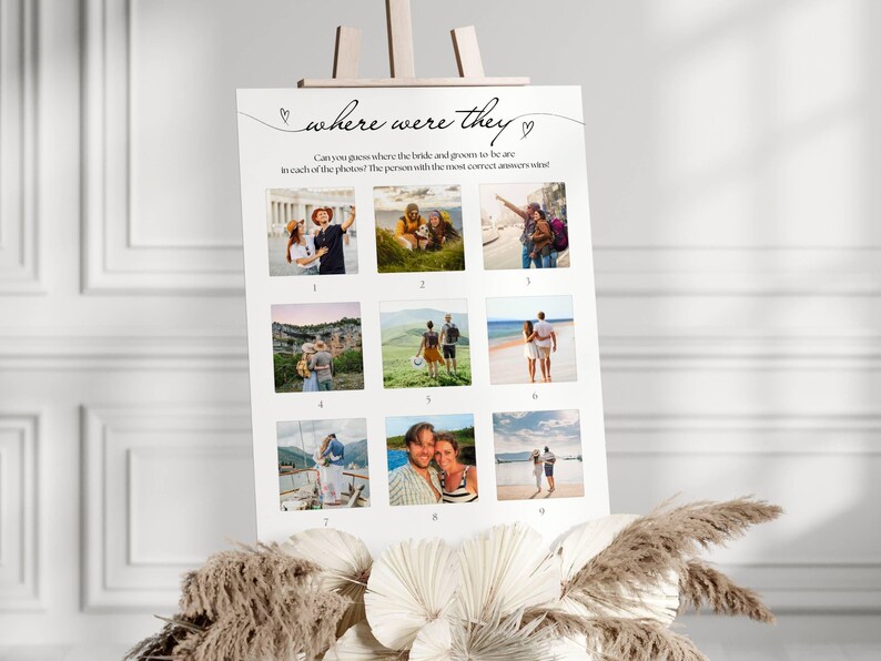 May include: A white sign on an easel with the text "where were they" and nine small photos. The sign is decorated with hearts and asks guests to guess the locations in the photos. The sign is decorated with dried flowers.