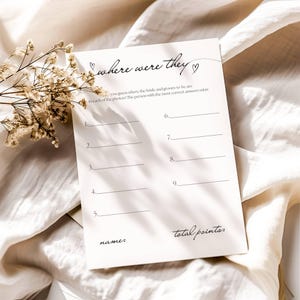 May include: A cream-colored wedding game card with the text "where were they" in a handwritten font. The card has lines for answers and the phrases "name:" and "total points:" at the bottom. A sprig of dried flowers rests on the left.