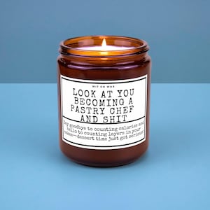 May include: A lit brown glass candle jar with a white label. The label reads "LOOK AT YOU BECOMING A PASTRY CHEF AND SHIT." The candle's text also says "Say goodbye to counting calories and hello to counting layers in your cakes-dessert time just got serious!"