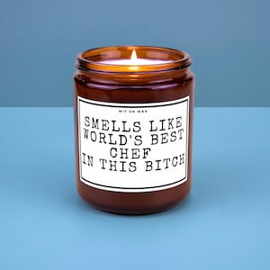 May include: A lit, brown glass jar candle with a white label. The label reads "Smells Like World's Best Chef In This Bitch" in black text. The candle sits on a blue surface.