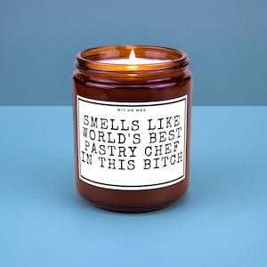 May include: A lit, brown glass jar candle with a white label. The label reads "Smells Like World's Best Pastry Chef In This Bitch". The candle sits on a blue surface.