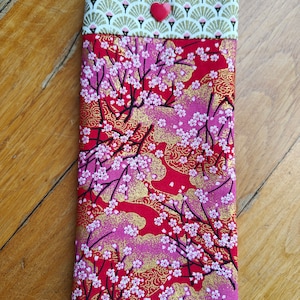May include: A rectangular fabric case with a red floral pattern featuring white blossoms and gold accents. The top section has a fan-shaped design in gold and white, secured with a red heart-shaped button. The case is likely for glasses or a phone.