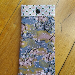 May include: A rectangular eyeglasses case with a floral pattern. The case features a design of cherry blossoms on a gray and pink background with gold accents. The top flap is a pink and white fan pattern, secured by a black button.