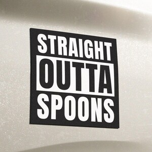 May include: A black and white rectangular bumper sticker with the text "STRAIGHT OUTTA SPOONS" in a bold, sans-serif font. The sticker is affixed to the back of a white vehicle. The vehicle has a red tail light and a 4x4 emblem.