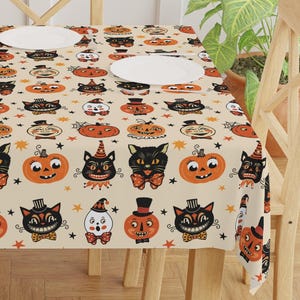 May include: A beige Halloween-themed tablecloth featuring a repeating pattern of black cats, pumpkins, and ghosts. The design includes orange, black, and white elements, with stars scattered throughout. The tablecloth is set on a wooden table with three white plates.