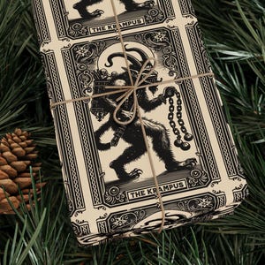 May include: Wrapped gift with a black and cream Krampus design, tied with twine, set against a backdrop of pine needles and pine cones. The gift wrap features a detailed illustration of Krampus holding chains.