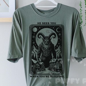 May include: Sage green t-shirt featuring a black and white illustration of a horned creature with the text "HE SEES YOU WHEN YOU'RE SLEEPING". The graphic includes a full moon, mountains, and houses.