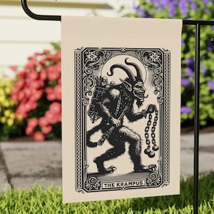 May include: A beige garden flag with a black and white illustration of a horned creature with a sack on its back and chains in its hand. The illustration is surrounded by a decorative frame and the text "THE KRAMPUS" is at the bottom.