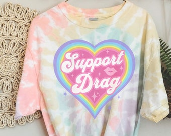 May include: A tie-dye t-shirt featuring a rainbow heart with the words "Support Drag" in a retro font. The heart has a pink background and a rainbow outline.