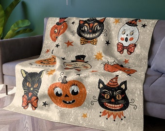May include: A cream-colored throw blanket with a Halloween theme. The blanket features a repeating pattern of black cats, pumpkins, and ghosts with stars and other festive details.