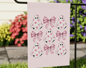 May include: A pink garden flag with a pattern of white ghosts with pink flowers and pink bows. The ghosts are smiling and have two eyes.