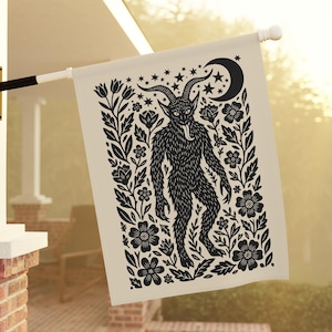 May include: A cream-colored outdoor flag featuring a black illustration of a horned creature with a long tongue, surrounded by floral and star designs, and a crescent moon. The flag is hanging outside.