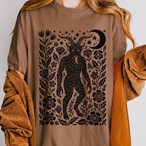 May include: Brown t-shirt featuring a black graphic of a horned creature with a long tongue, surrounded by flowers, stars, and a crescent moon. The shirt is a neutral color and has short sleeves.