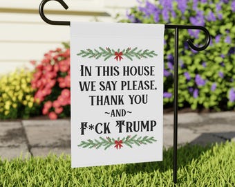May include: White garden flag with black text that reads "IN THIS HOUSE WE SAY PLEASE, THANK YOU ~AND~ F*CK TRUMP." The text is framed by green sprigs and red bows. The flag is displayed on a black metal stand in a grassy yard.