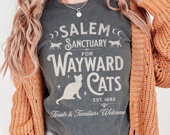 May include: A gray t-shirt with a white graphic that says "Salem Sanctuary for Wayward Cats Est. 1692 Ferals & Familiars Welcome". The graphic also includes a crescent moon, stars, and a cat silhouette.