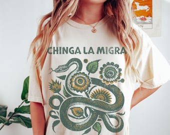 May include: Cream-colored t-shirt featuring a green snake intertwined with green and yellow flowers. The text "CHINGA LA MIGRA" is printed above the snake design. The shirt has short sleeves and a relaxed fit.