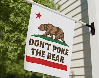 May include: A white flag with the California state flag design. It features a brown bear, a red star, and the text "DON'T POKE THE BEAR" in dark brown. A red stripe is at the bottom.
