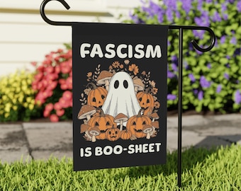 May include: Black garden flag with white text reading "FASCISM IS BOO-SHEET". The flag shows a cartoon ghost, orange pumpkins, and mushrooms. It's displayed on a black metal stand in a garden with grass and flowers.