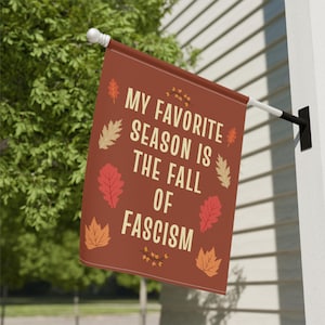 May include: A rectangular brown flag with the text "MY FAVORITE SEASON IS THE FALL OF FASCISM" in white. The flag is decorated with fall leaves in various colors. The flag is hanging outside.