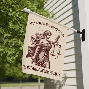 May include: A beige flag with the words "WHEN INJUSTICE BECOMES LAW RESISTANCE BECOMES DUTY" and an illustration of a woman holding scales and a sword. The flag is hanging outside on a white wall.