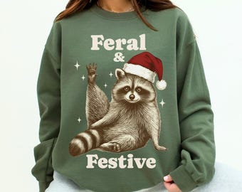 May include: Green sweatshirt featuring the text "Feral & Festive" and a raccoon wearing a Santa hat. The raccoon is sitting and waving. White stars are also included in the design.
