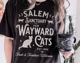 May include: Black t-shirt with white text that reads "Salem Sanctuary for Wayward Cats Est. 1692 Ferals & Familiars Welcome". The text is in a vintage style font. The shirt features a black cat silhouette and a crescent moon.