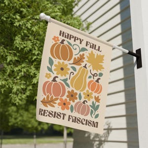 May include: A beige flag with the words "HAPPY FALL" and "RESIST FASCISM" in brown text. The flag features illustrations of pumpkins, flowers, and leaves in orange, brown, and yellow. The flag is hanging on a white wall.