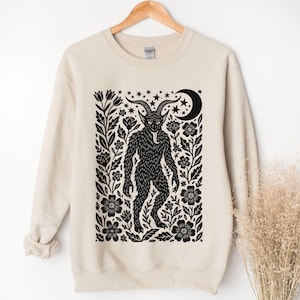 May include: Beige sweatshirt featuring a black illustration of a horned creature with a long tongue, surrounded by floral and star designs. The design includes a crescent moon and is printed on a crew neck sweatshirt.