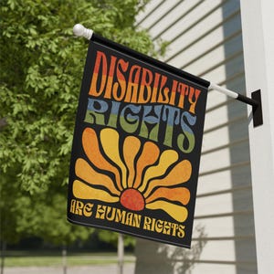May include: A black flag with the words "DISABILITY RIGHTS ARE HUMAN RIGHTS" in a retro font. The flag features a colorful sunburst design in shades of orange, yellow, and red. The flag is mounted on a white and black pole.