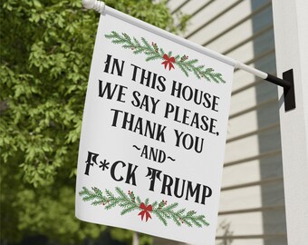 May include: White house flag with the text "IN THIS HOUSE WE SAY PLEASE, THANK YOU ~AND~ F*CK TRUMP" in black. The flag is decorated with green garland and red bows. The flag is hanging outside.