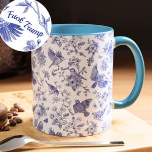 May include: White ceramic mug with a blue interior and handle. The mug features a blue and white floral and bird pattern with the text "Fuck Trump" repeated throughout the design. The mug is sitting on a wooden surface.