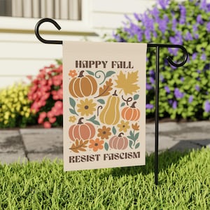 May include: A beige garden flag with the words "HAPPY FALL" and "RESIST FASCISM" in brown text. The flag features illustrations of pumpkins, flowers, and leaves in shades of orange, brown, and yellow. The flag is displayed on a black metal stand.