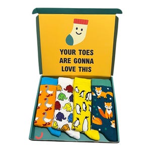 May include: A gift box containing four pairs of patterned socks. Designs include geese, elephants, penguins, and foxes. The box displays the text "YOUR TOES ARE GONNA LOVE THIS" and a smiling sock illustration.