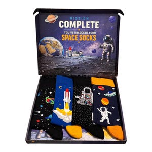 May include: A gift box containing four pairs of space-themed socks. The box lid features a space scene with an astronaut, Earth, and the text MISSION COMPLETE YOU'VE UNLOCKED YOUR SPACE SOCKS. The socks feature patterns of rockets, astronauts, and planets.