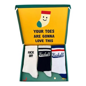 May include: A gift box containing three pairs of socks. The box has a yellow interior with the text "YOUR TOES ARE GONNA LOVE THIS" and a cartoon sock. The socks are white, navy, and white with the words "FUCK OFF" and "Fuckoff" printed on them.