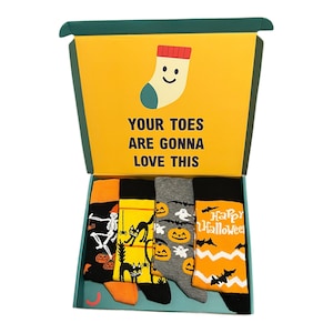 May include: A gift box containing four pairs of Halloween-themed socks. The socks feature designs of skeletons, black cats, ghosts, pumpkins, and bats. The box has the text "YOUR TOES ARE GONNA LOVE THIS" and a cartoon sock.