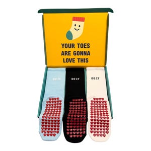 May include: A gift box containing three pairs of socks. The yellow box features a cartoon sock and the text "YOUR TOES ARE GONNA LOVE THIS". The socks are light blue, black, and white, each with red heart-shaped grips and the text "DO IT".
