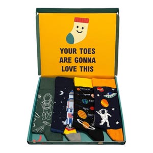 May include: A gift box containing four pairs of space-themed socks. The box lid features a yellow background with a smiling sock icon and the text "YOUR TOES ARE GONNA LOVE THIS". The socks feature designs of an astronaut, a rocket, planets, and stars.