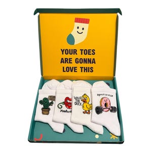 May include: A set of four white crew socks with colourful graphic prints inside a green and yellow gift box. The box lid features a smiling sock illustration and the text "YOUR TOES ARE GONNA LOVE THIS". The socks feature a cactus, lips, a duck, and a donut.
