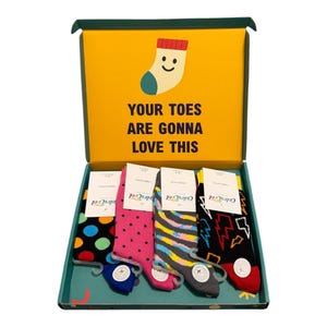 May include: A gift box containing four pairs of patterned socks. The yellow interior lid features a cartoon sock and the text YOUR TOES ARE GONNA LOVE THIS. The socks feature polka dots, stripes, and lightning bolt designs in vibrant colours.
