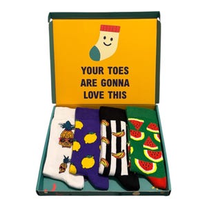May include: A gift box containing four pairs of crew socks with fruit patterns. The box lid features a yellow background with a smiling sock icon and the text YOUR TOES ARE GONNA LOVE THIS. The socks feature pineapple, lemon, banana, and watermelon prints.