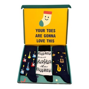May include: A gift box containing three pairs of socks. The left pair is dark blue with colourful electric guitars. The middle pair is white with black musical notes and text. The right pair is dark blue with various musical instruments.