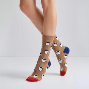 May include: Tan socks featuring a pattern of white coffee cups with black coffee. The heels are blue, and the toes are red. The socks are knee-high.