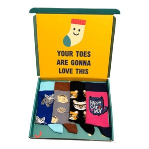 May include: A gift box containing four pairs of patterned socks with cat designs. The box has a yellow lid with the text "YOUR TOES ARE GONNA LOVE THIS." The socks include a blue pair with cat illustrations, a grey pair with cat faces, a black pair with cats wearing glasses, and a pink pair with a cat graphic.