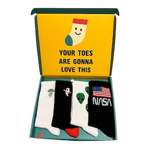 May include: A gift box containing four pairs of socks. The socks are white and black with designs such as a mushroom, an astronaut, green aliens, and a NASA logo with a US flag. The box has a yellow interior with the text "YOUR TOES ARE GONNA LOVE THIS".