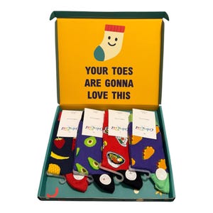 May include: A gift box containing four pairs of colourful patterned socks with food designs including strawberries, avocados, ramen bowls, and tacos. The box lid features a yellow background with a sock illustration and the text YOUR TOES ARE GONNA LOVE THIS.