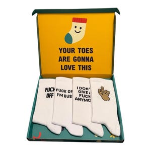 May include: A set of four white crew socks inside a yellow and green cardboard box. The box lid features a sock illustration and the text "YOUR TOES ARE GONNA LOVE THIS". The socks display black text and a brown hand graphic showing a middle finger.
