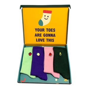 May include: A gift box containing four pairs of ribbed socks in light green, purple, pink, and dark green. Each pair features a small embroidered fruit icon: an apple, lemon, watermelon slice, and avocado. The box lid reads, YOUR TOES ARE GONNA LOVE THIS.